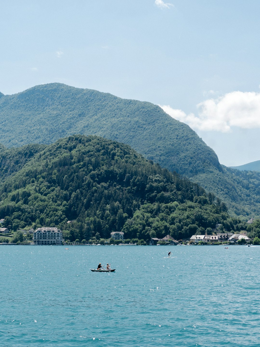 Two people kayak on a blue lake near forested mountains.