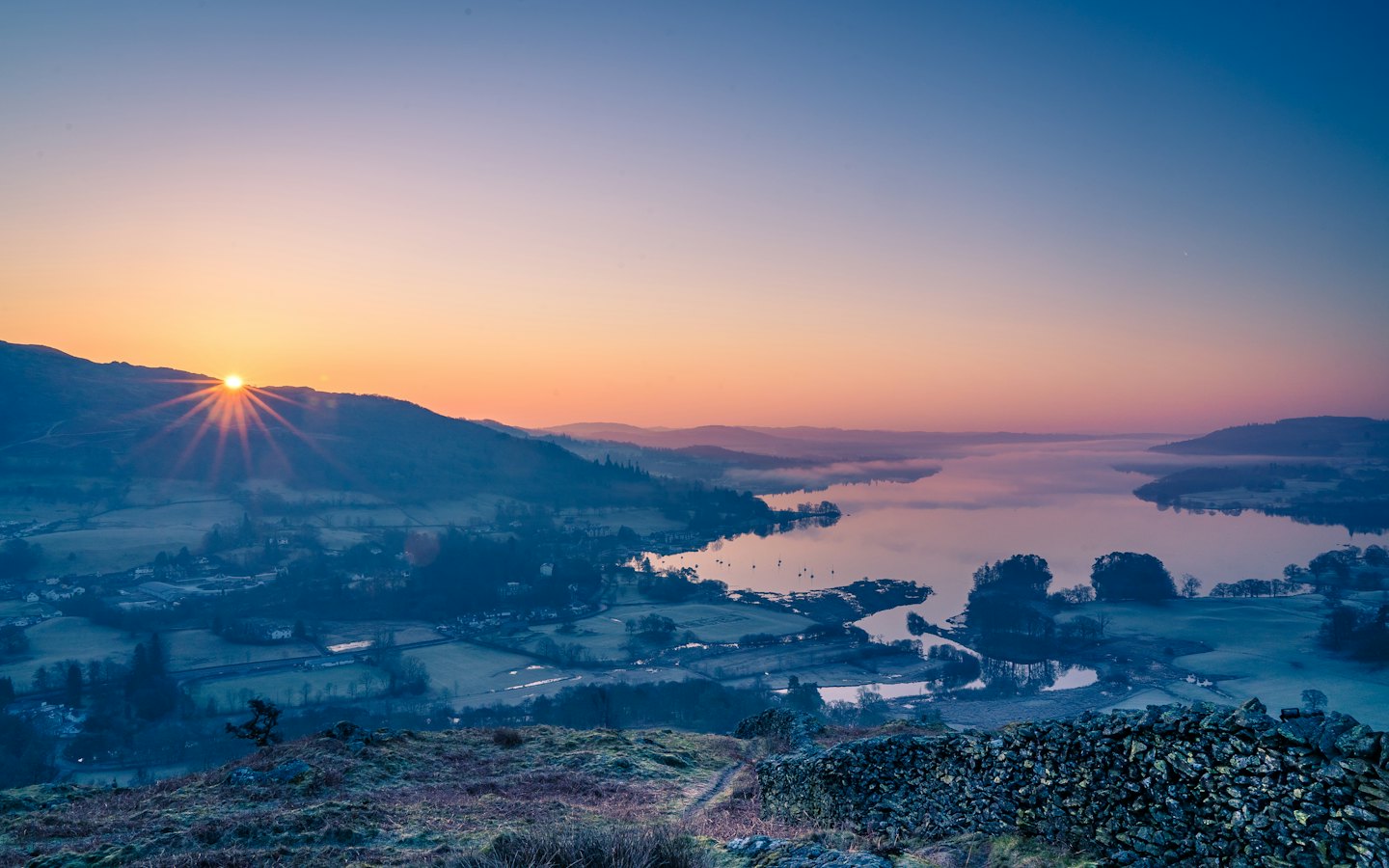 Sunrise lights a misty lake surrounded by jagged mountain peaks.